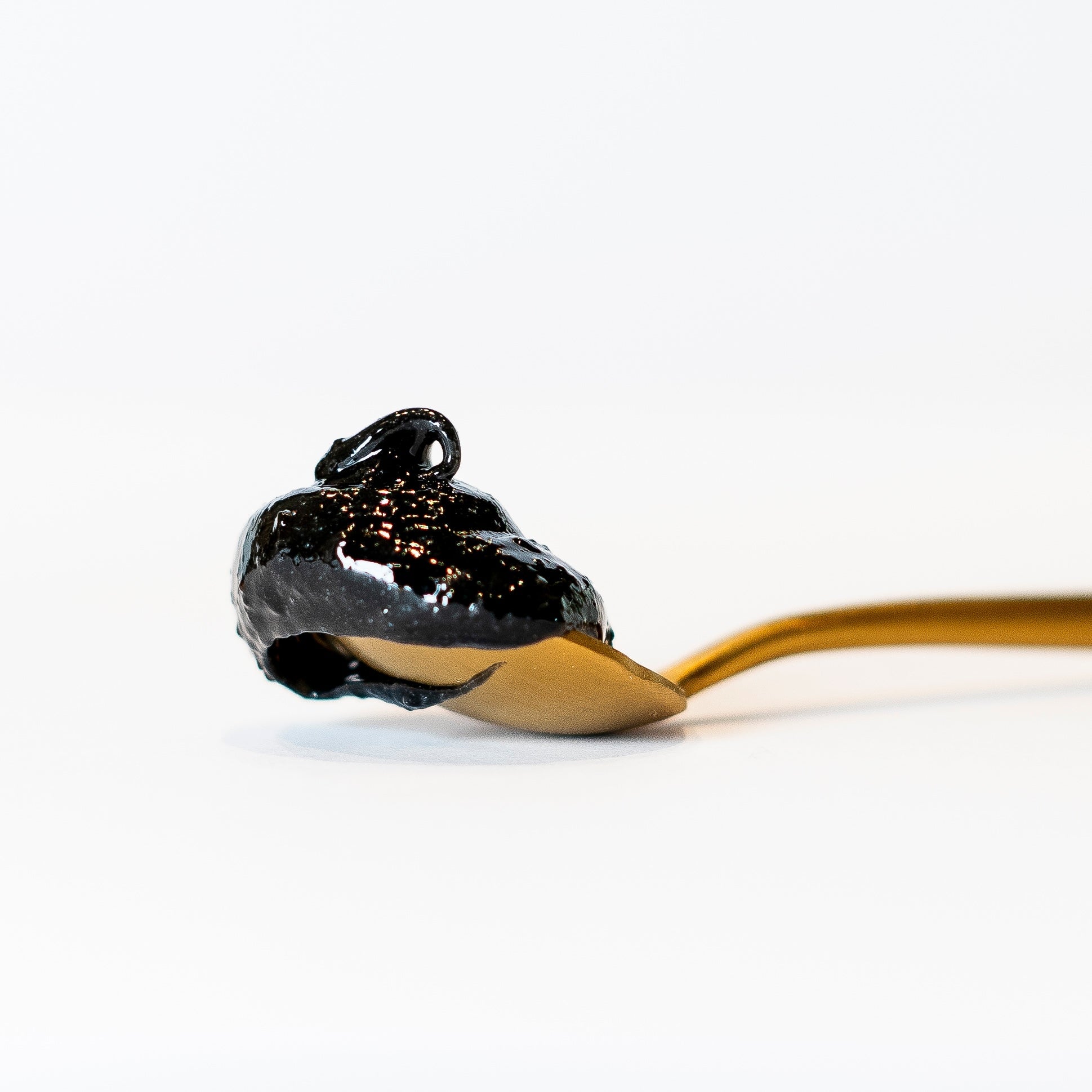 Ein Messinglöffel mit schwarzer, glänzender Masse drauf, liegt auf hellem Untergrund.
