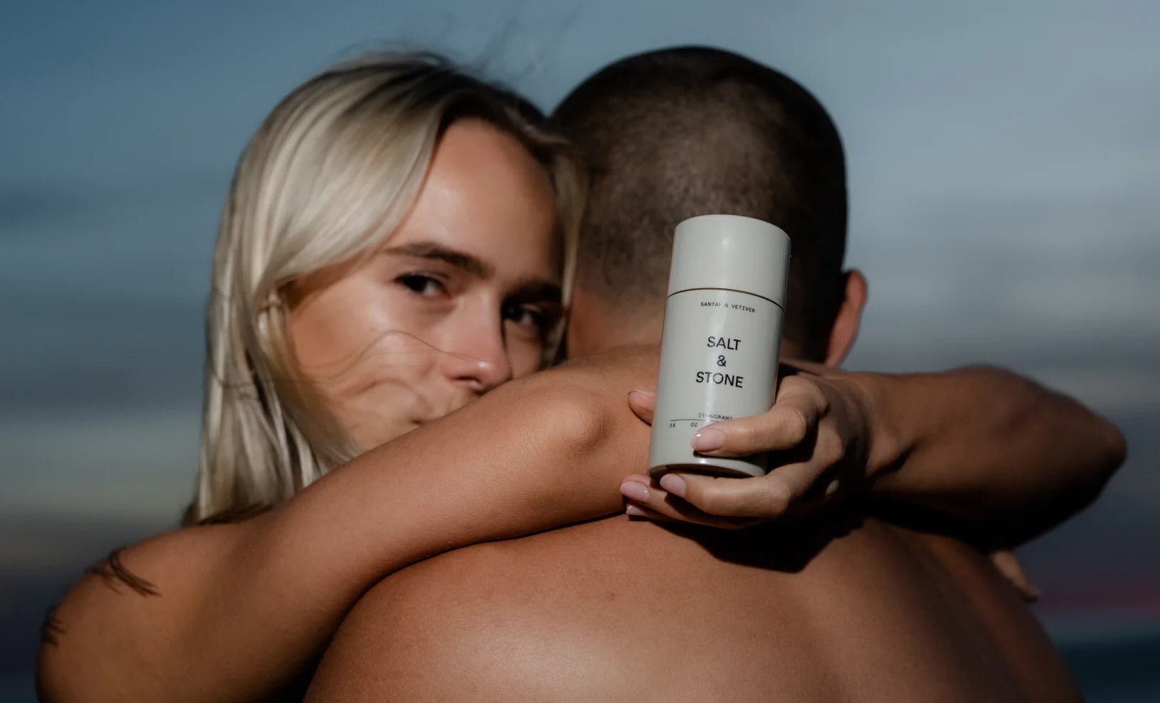 Blonde Frau umarmt einen Mann und hält dabei ein Salt & Stone Deodorant in der Hand.