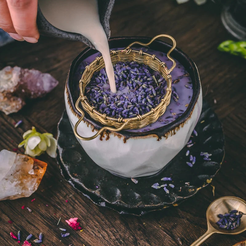 Anima Mundis Wooven Brass Large Tea Strainer - großes Teesieb gefüllt mit Lavendel wird mit milchiger Flüssigkeit aufgegossen in einer großen Tasse, steht auf einem Holztisch, dekoriert mit Edelsteinen und Blütenblättern.