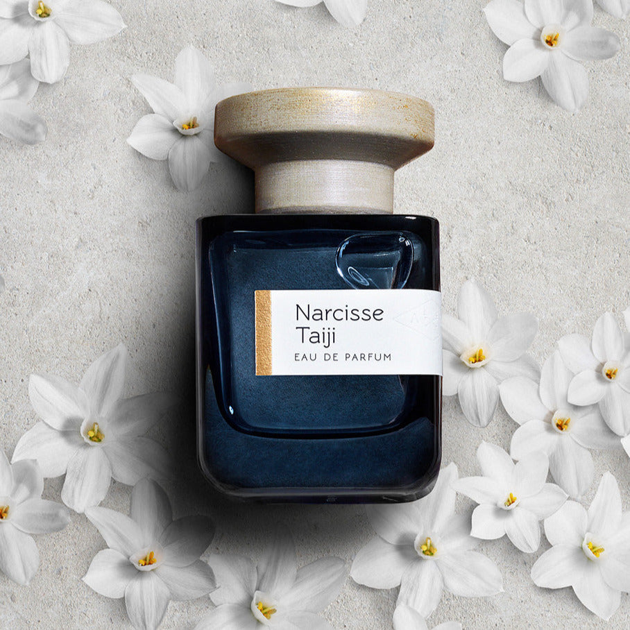 Parfumflasche Narcisse Taiji liegend auf weißem Sand mit Blüten von Narcissen. North Glow