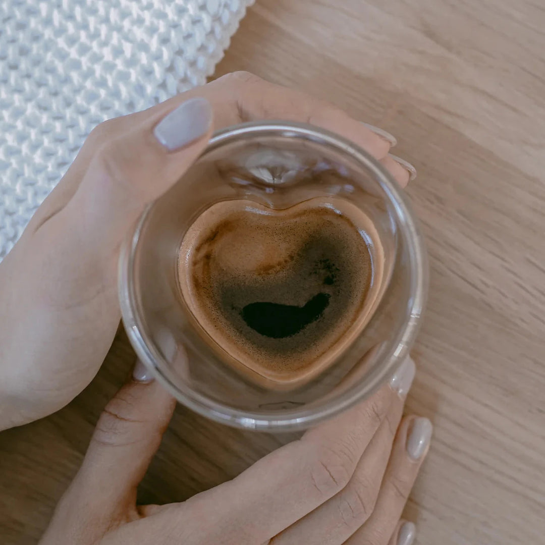Herzförmiges Glas mit Kaffee wir von zwei Händen gehalten, stehend auf einem Holztisch.