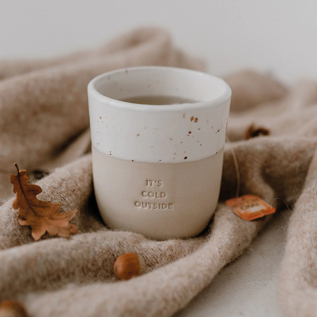 Kaffeebecher mit Prägung "it's cold outside" steht auf einer hellbraunen Decke umgeben von einem Eichenblatt und einer Eichel. North Glow