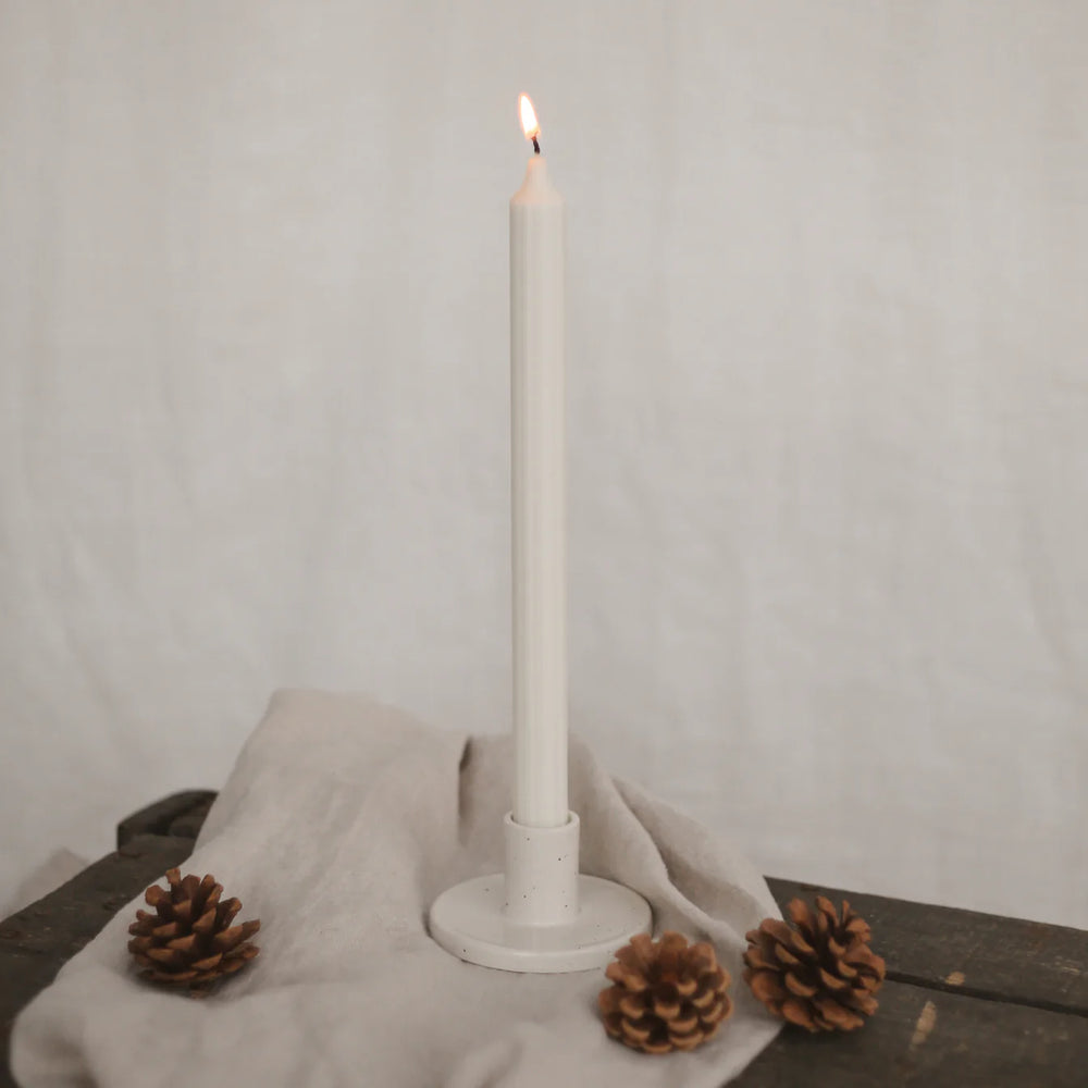 Angezündete weiße Stabkerze von Eulenschnitt in einem Kerzenhalten auf einem beigen Leinentuch, daneben 3 Tannenzapfen.
