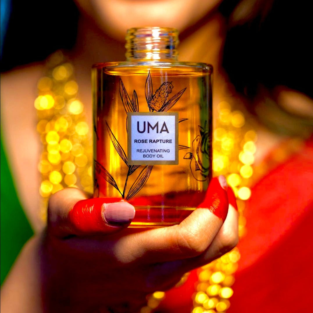 Frau verschwommen im Hintergrund mit bunter Kleidung und vielen Goldketten hält in den Vordergrund die UMA Rose Rapture Rejuvenationg Body Oil Flasche. North Glow