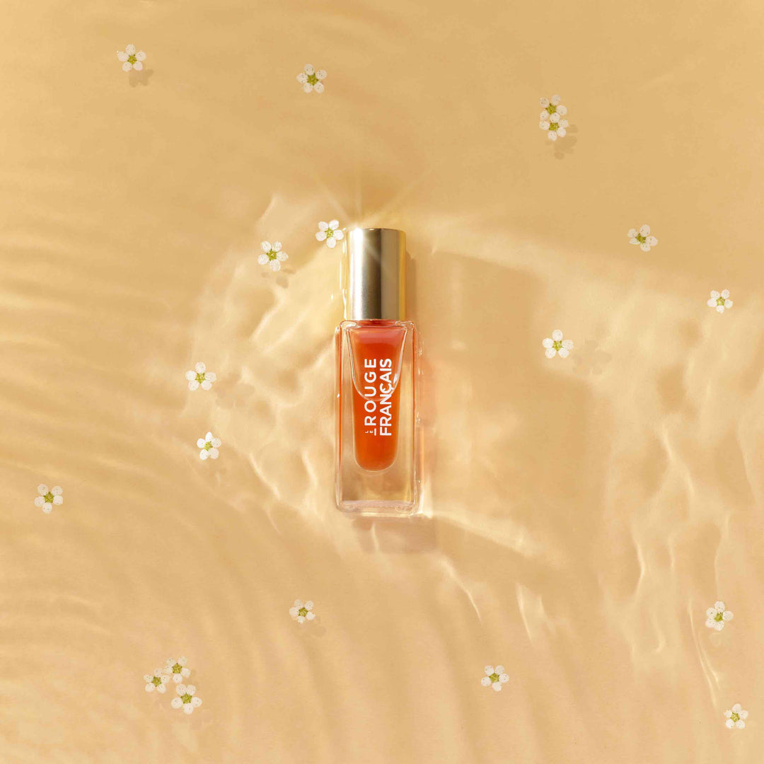 Lippenöl Glasflasche von Le Rouge Francais Orange Persephone liegt im seichten Wasser im Sand umringt von Blütenblättern. North Glow