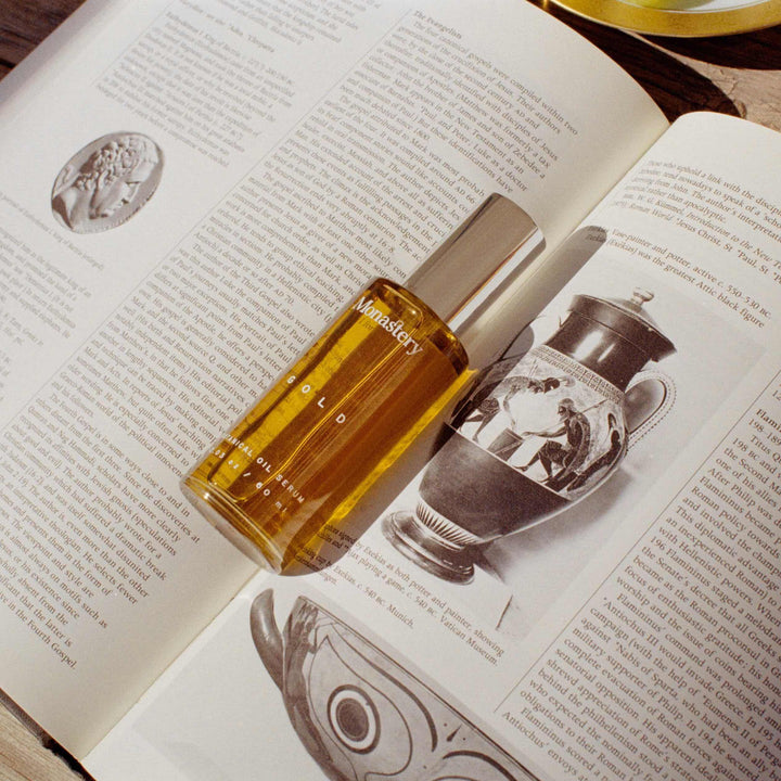 Monastery Gold Serum Glasflasche mit silbernem Deckel liegt auf aufgeschlagenem Buch auf einem Holztisch.