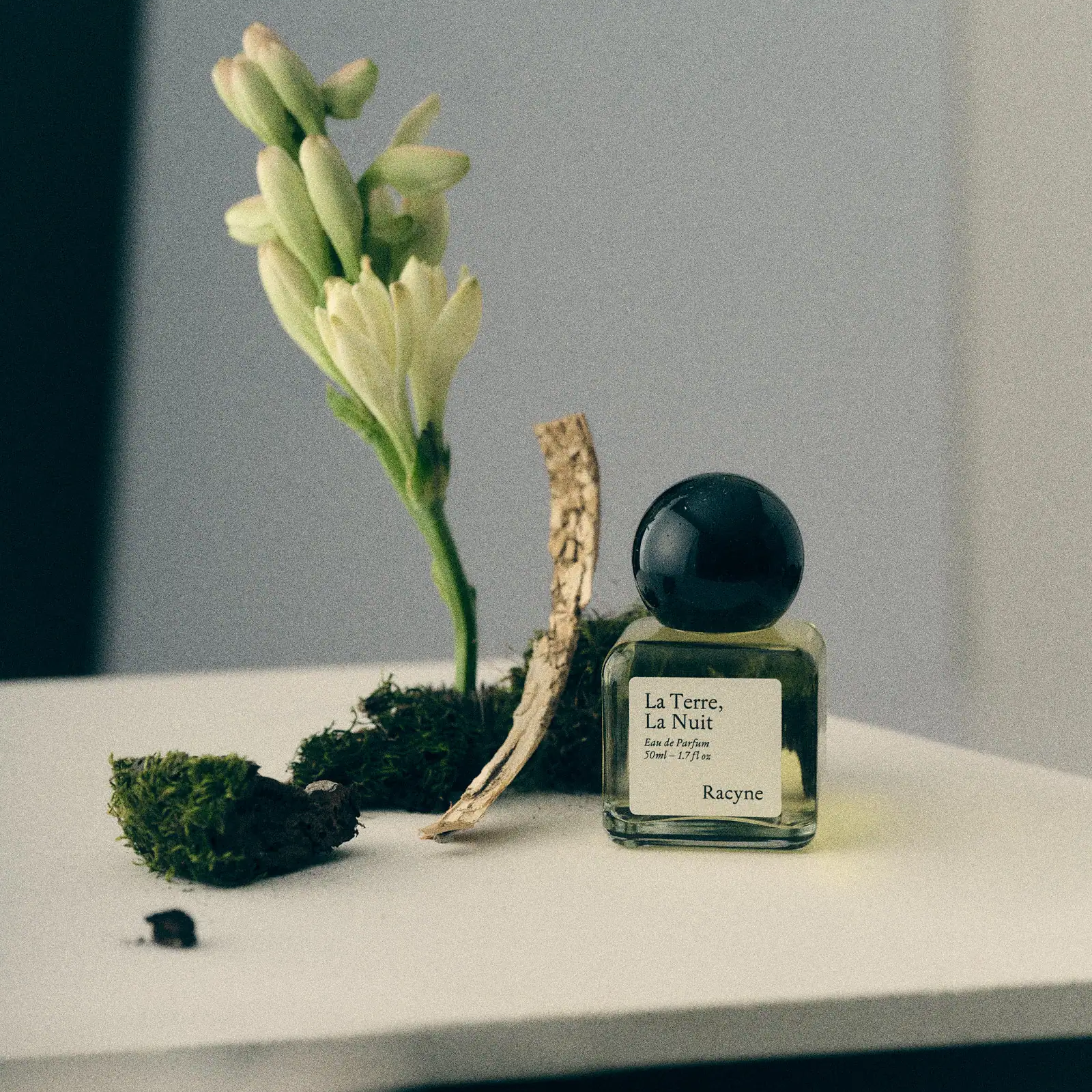 Racyne La Terre, La Nuit 50 ml Flakon mit schwarzem Kugelverschluss neben Moos, heller Blüte und Holz.