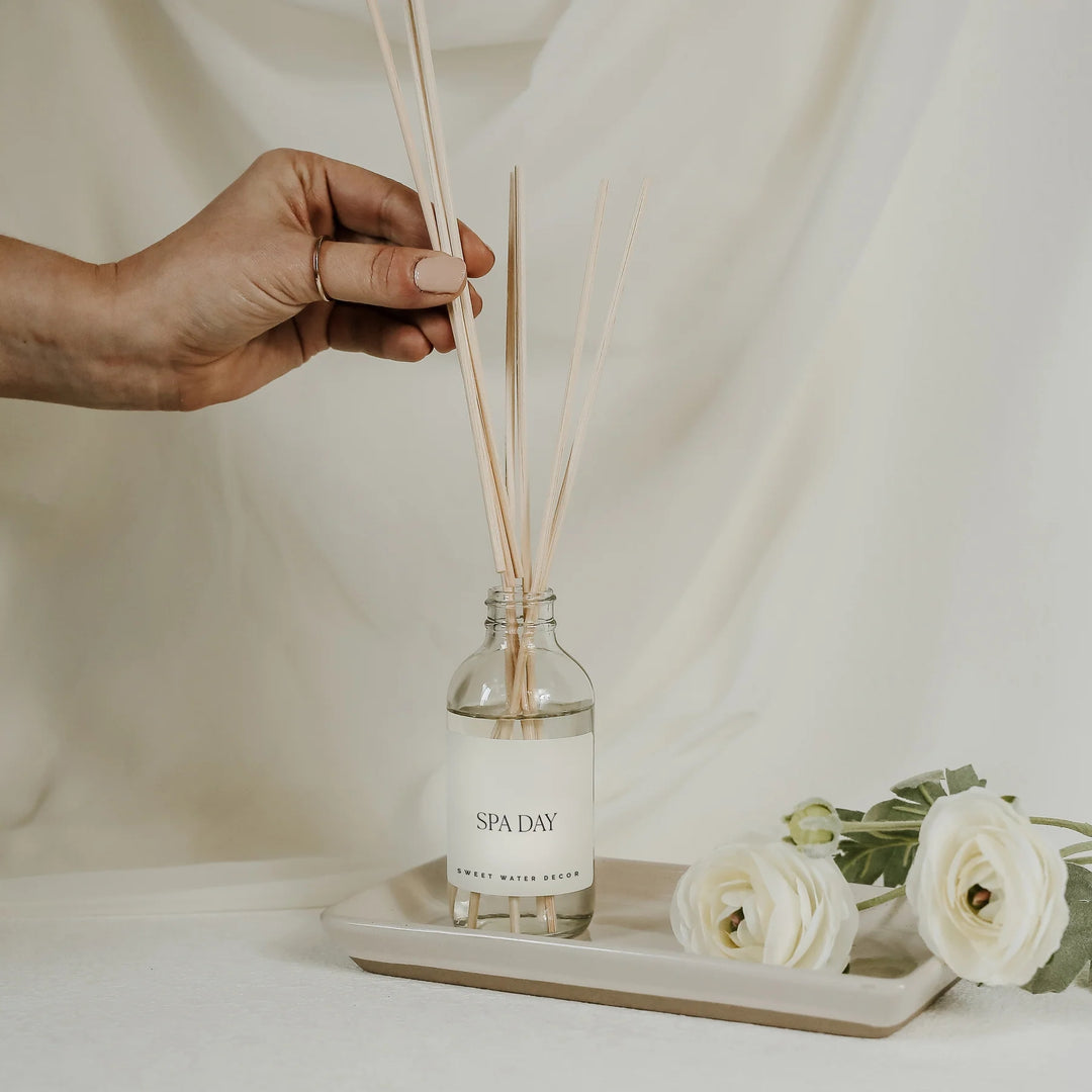 Hand hält Schilfrohrstäbchen in eine Flasche Sweet Water Decor Diffuser Spa Day, die auf einem Tablett steht mit weißen Blumen vor hellem Hintergrund. North Glow