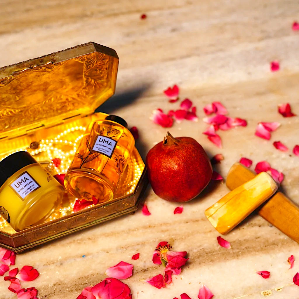 Flasche und Schraubglas des UMA Rose Rapture Body Balms und Oils liegen in einer goldenen Schachtel, welche auf einer Marmorplatte liegt, dekoriert mit Granatapfel und Blütenblättern. North Glow