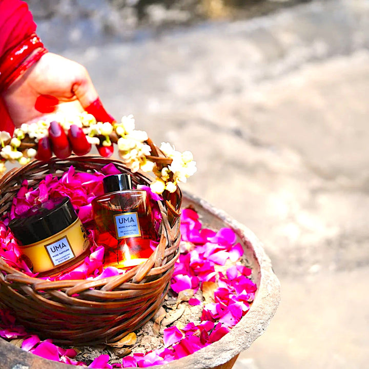 Hand mit roten Fingespitzen hält ein mit Blütenblättern gefülltes Weidenkörbchen in dem Flasche und Schraubglas des UMA Rose Rapture Body Balms und Oils liegen.