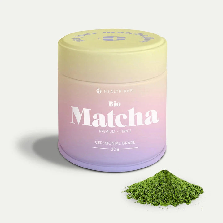 Bio Matcha von Health Bar in pastellfarbener Verpackung vor hellem Hintergrund, davor grünes Matcha-Pulver.