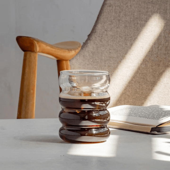 Mit Kaffee befülltes Bubble Glas Blanko von Eulenschnitt steht auf einem Tisch vor einem Stuhl und neben einem Buch.