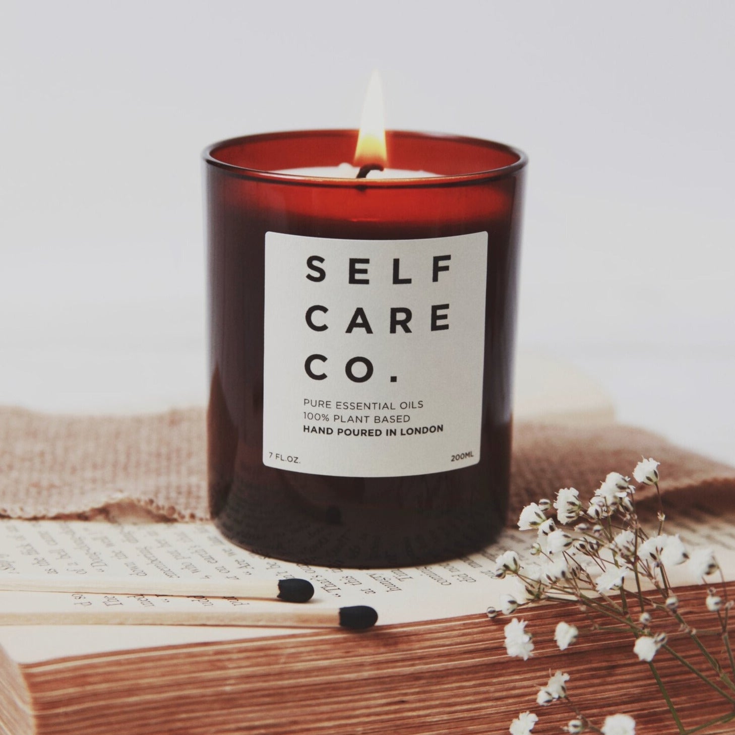 Braunes Kerzenglas mit Aufschrift Self Care Co. steht auf einem Buch, im Vordergund liegen zwei Streichhölzer
