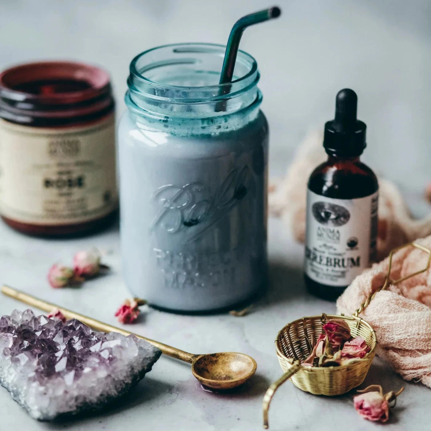 Anima Mundi Cerebrum Pipettenglasflasche steht neben einem hellblauem Getränk, davor Blütenblätter und ein Messinglöffel.