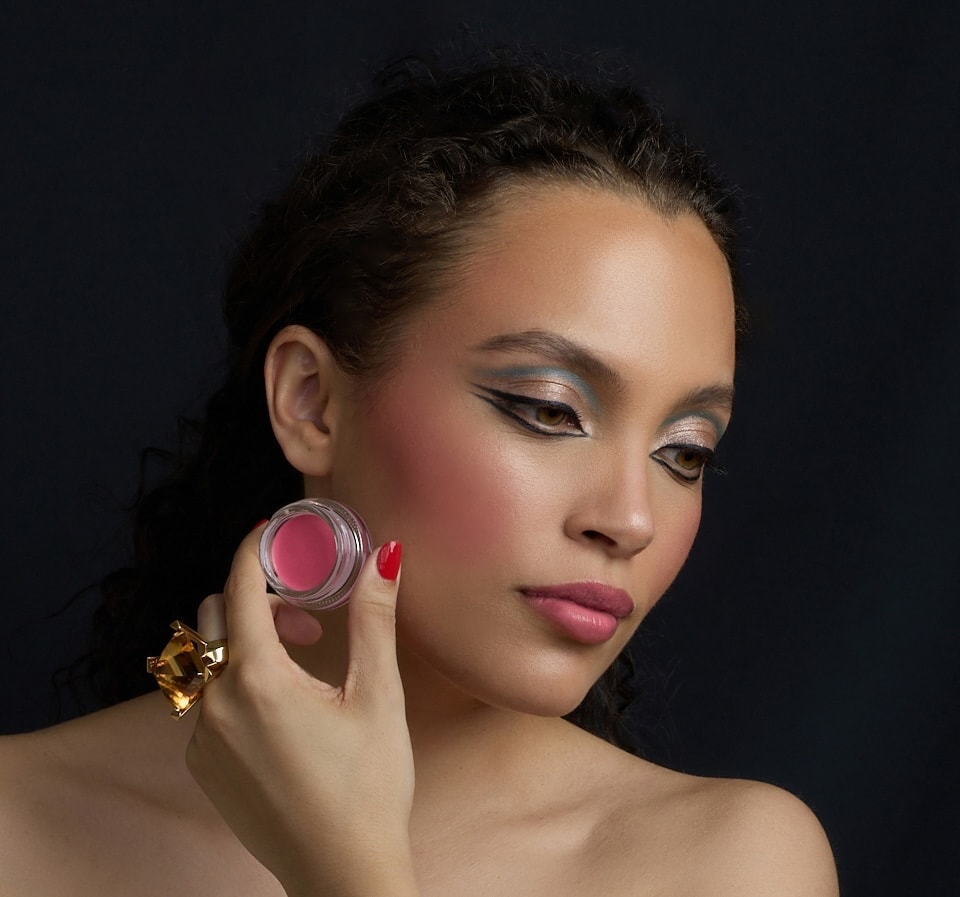 Eine Frau hält den Cream Blush CLEPATRE von Le Rouge Francais neben ihr Gesicht.