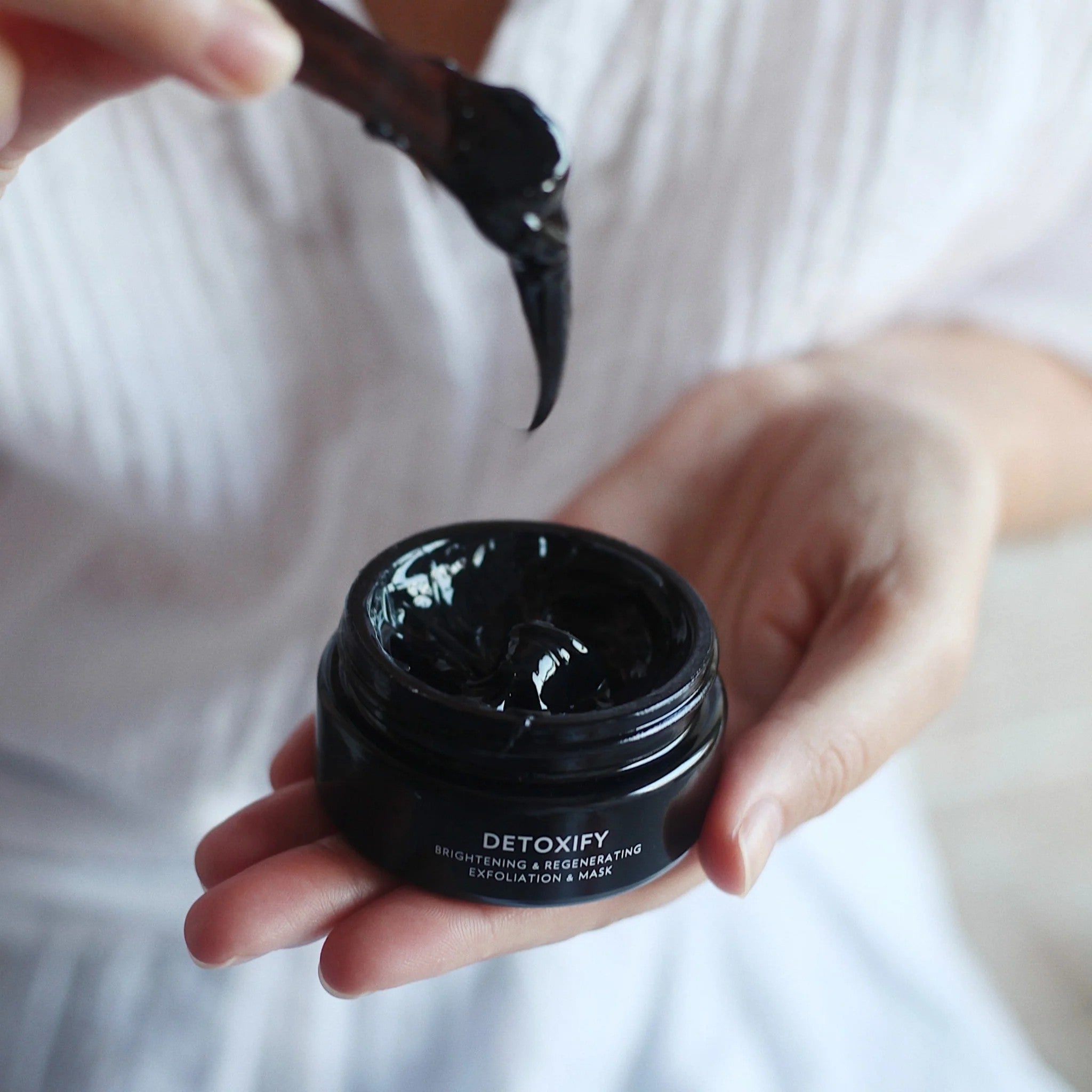 Offenes, Schwarzes Schraubglas Dafna's Detoxify Peeling Maske mit schwarzem, gelartigem Inhalt - wird umgerührt mit einem Spatel.