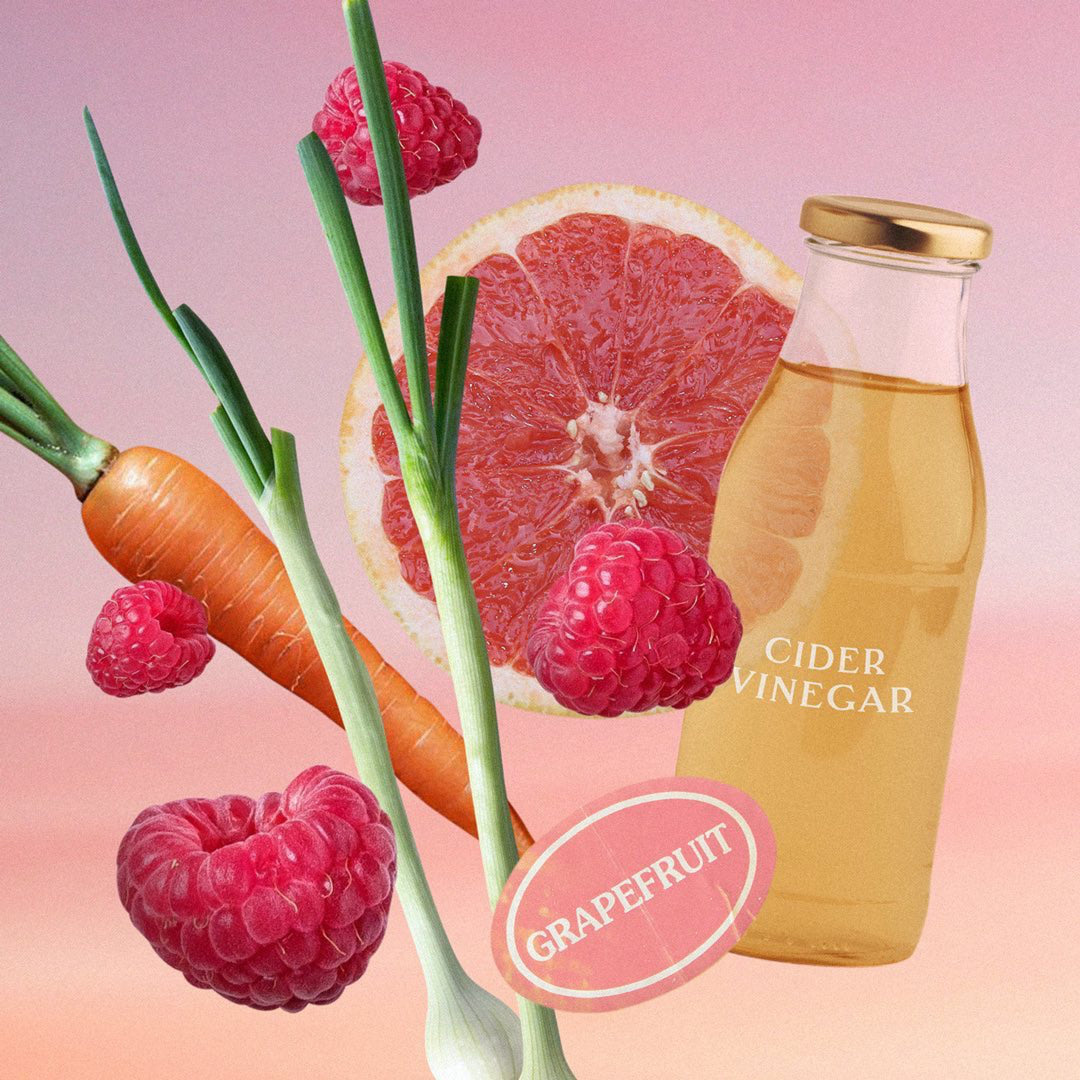 Vor rosafarbenem Hintergrund sind Himbeeren, eine aufgeschnittene Grapefruithälfte, Karotte, Frühlingszwiebeln und eine Flasche Cider Vinegar. North Glow