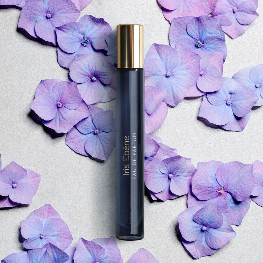 Längliches Parfumgefäß "Iris Ebéne" von Atelier Materi umgeben von Blüten.