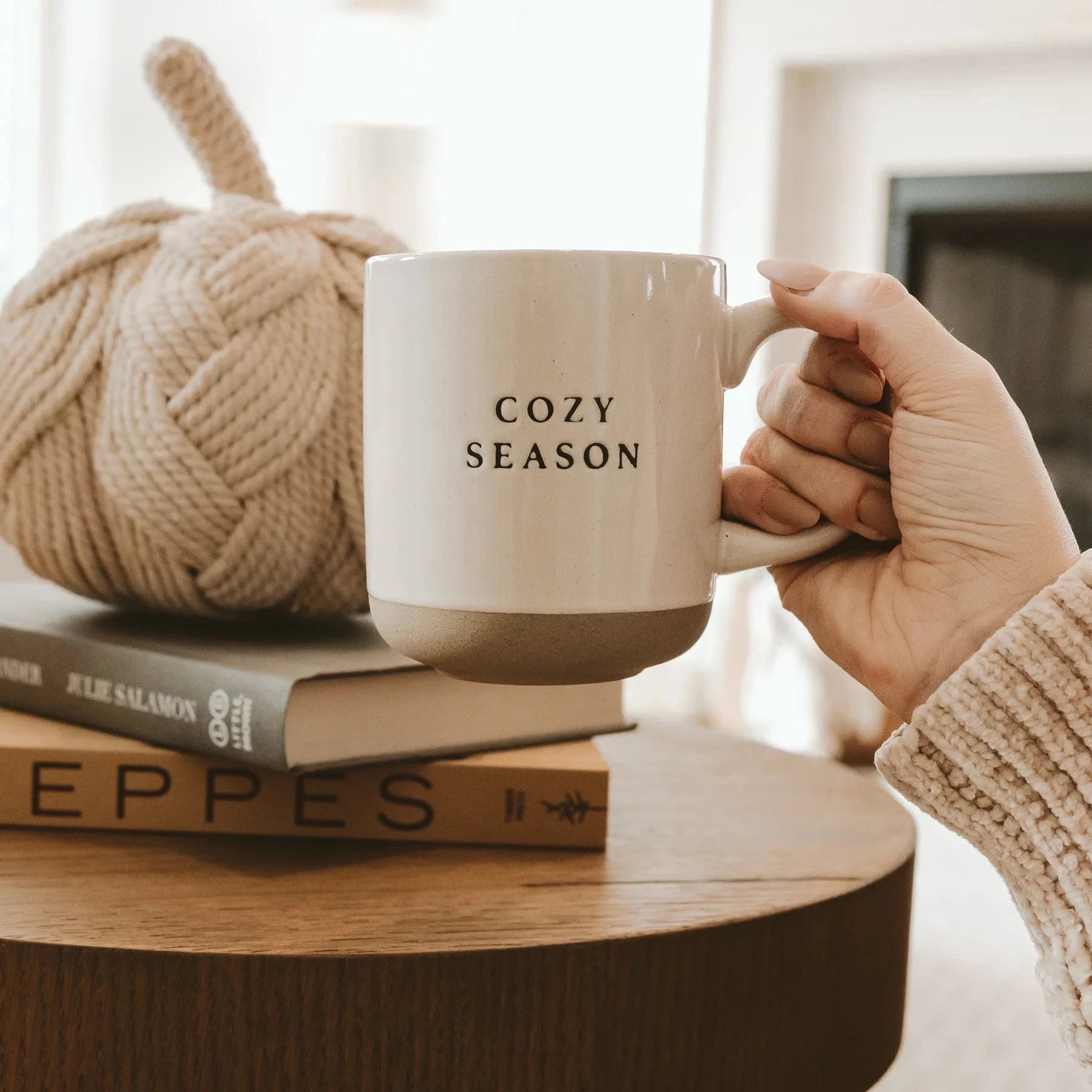 Eine Hand hält die Kaffeetasse Cozy Season von Sweet Water Decor über einem Holzsch fest.