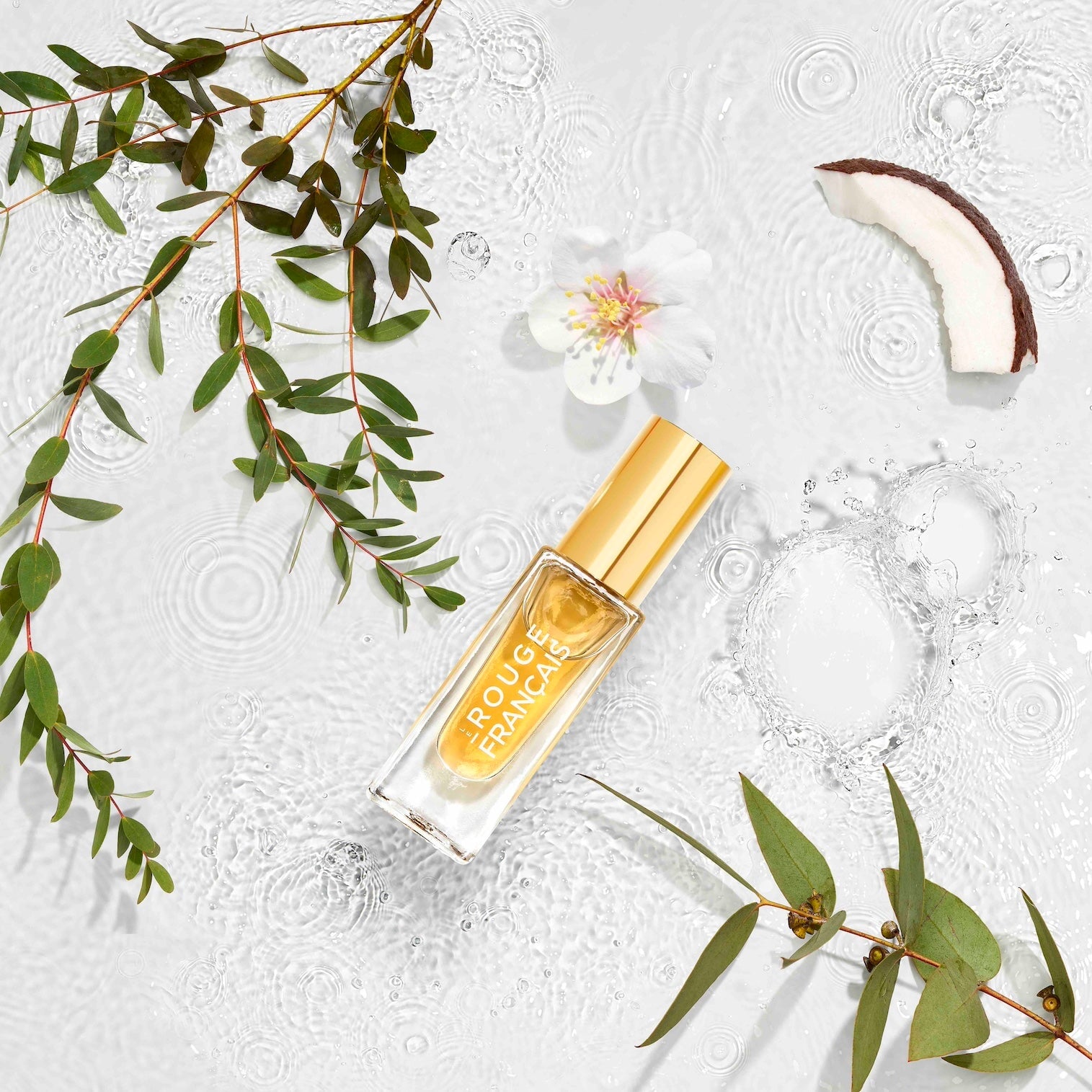 Glasflasche Le Nectar mit farblosen Lippenöl liegt im seichten Wasser mit einer weißen Blüte, einem Stück Kokosnuss und grünen Zweigen.