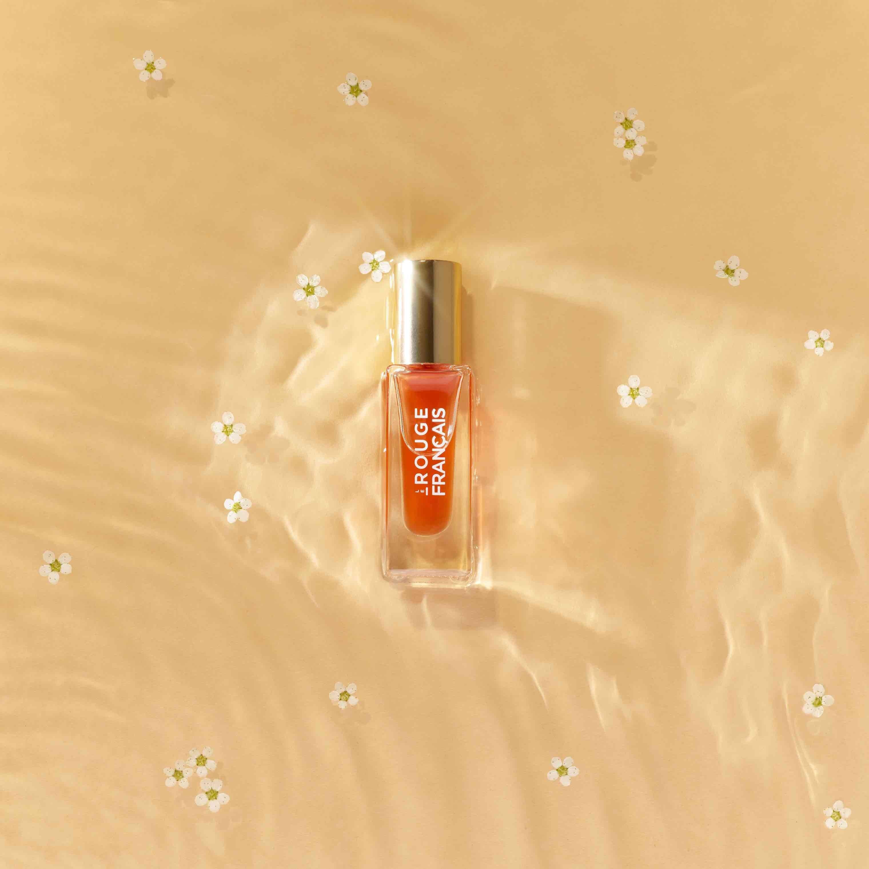 Lippenöl Glasflasche von Le Rouge Francais Orange Persephone liegt im seichten Wasser im Sand umringt von Blütenblättern.