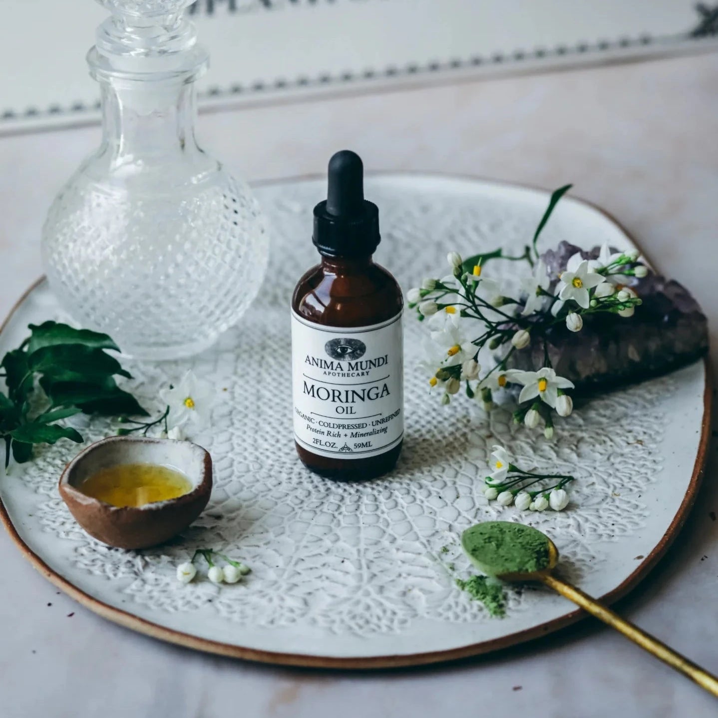 Glasflasche Moringa Oil von Anima Mundi, stehend, auf einem weißen, mit Blumen geprägtem Teller.
