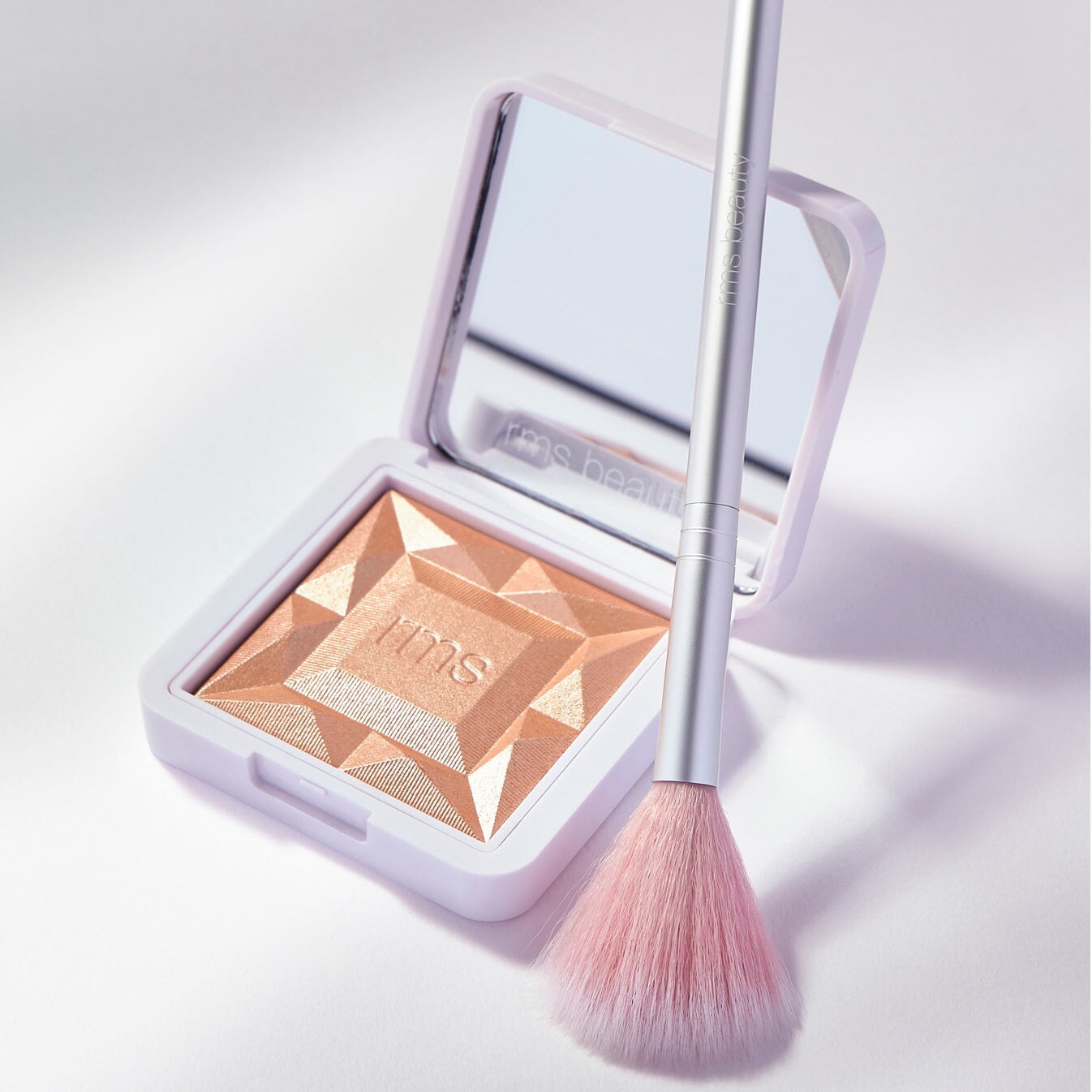 Skin2Skin Classic Fan Brush von RMS steht angelehnt an einer offenen RMS Highlighter Pudersdose.