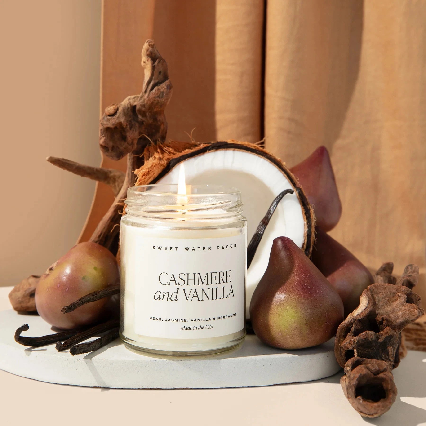 Brennende Sojawachskerze Cashmere and Vanilla steht zwischen Birnen, halber Kokosnuss, Vanilleschoten und Baumrinde.