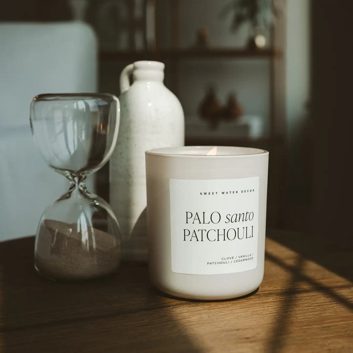 Duftkerze Palo santo Patchouli im Milchglas steht neben einer Sanduhr auf einem Holztisch.