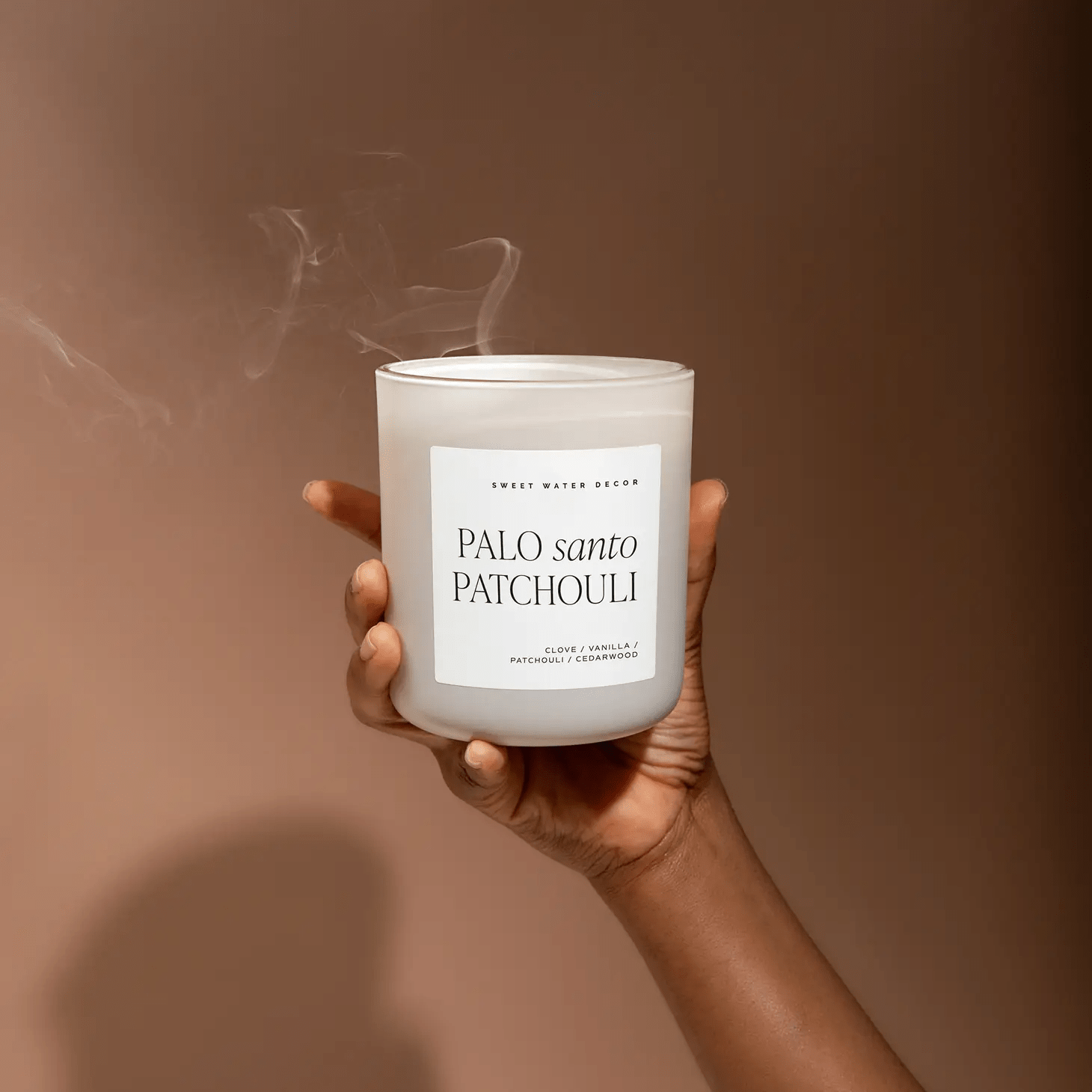 Eine Hand hälte eine leicht qualmende Duftkerze Palo santo Patchouli im Milchglas hoch.