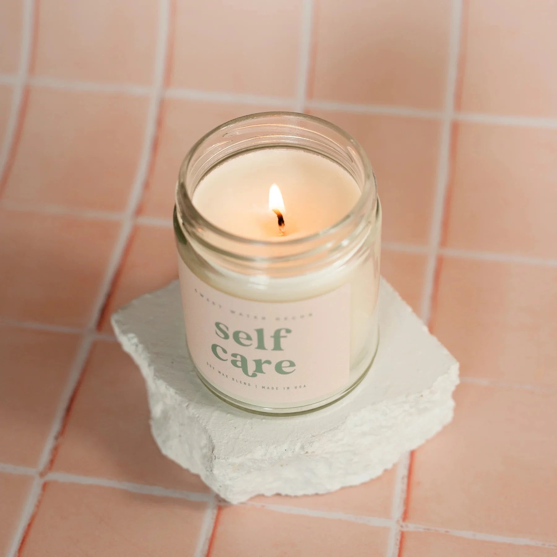 Brennende Sojawachskerze Self Care steht auf einem gefliestem Untergrund.
