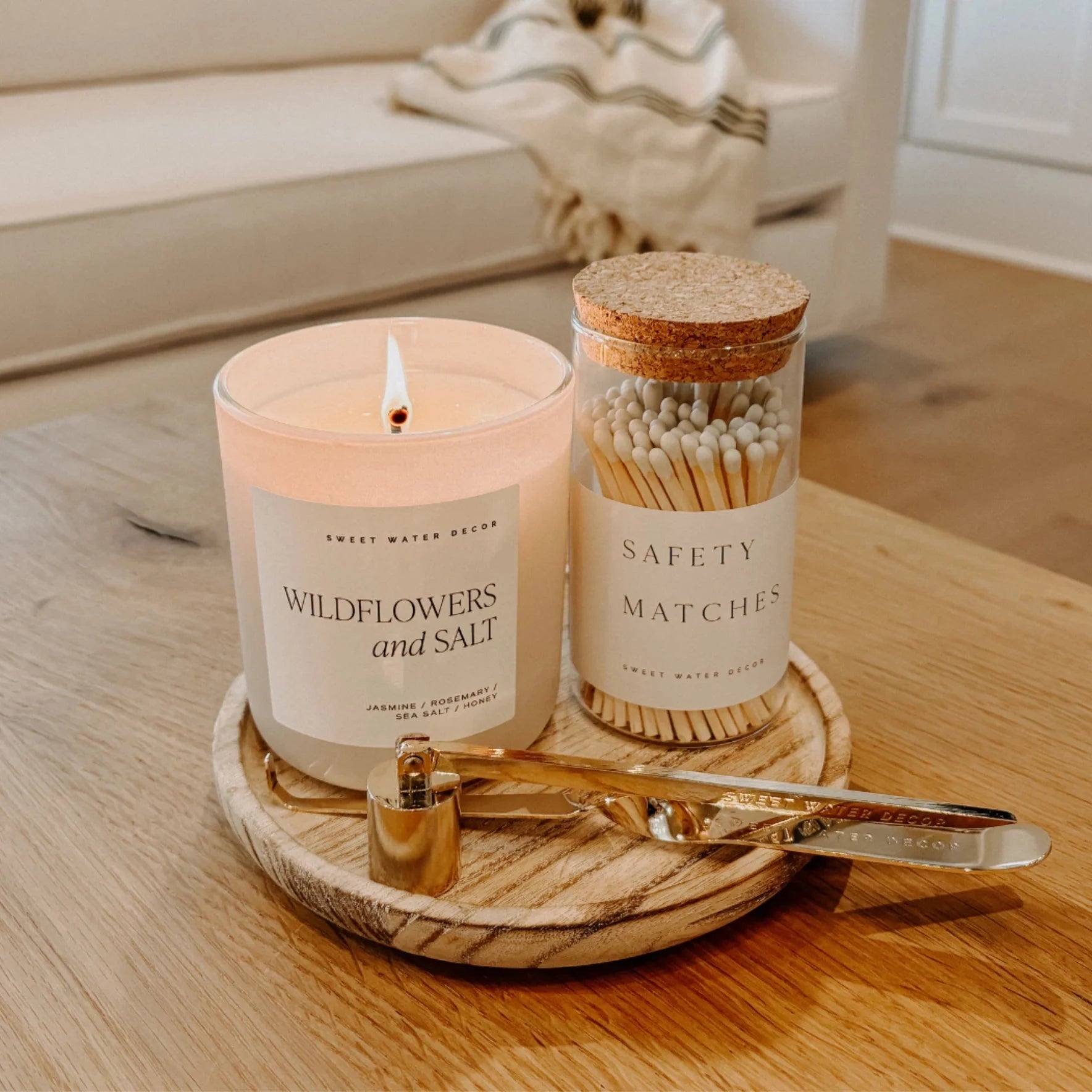 Duftkerze Wildflowers and Salt von Sweet Water Decor steht neben Streichhölzern auf einem Holzteller in einem Wohnzimmer.