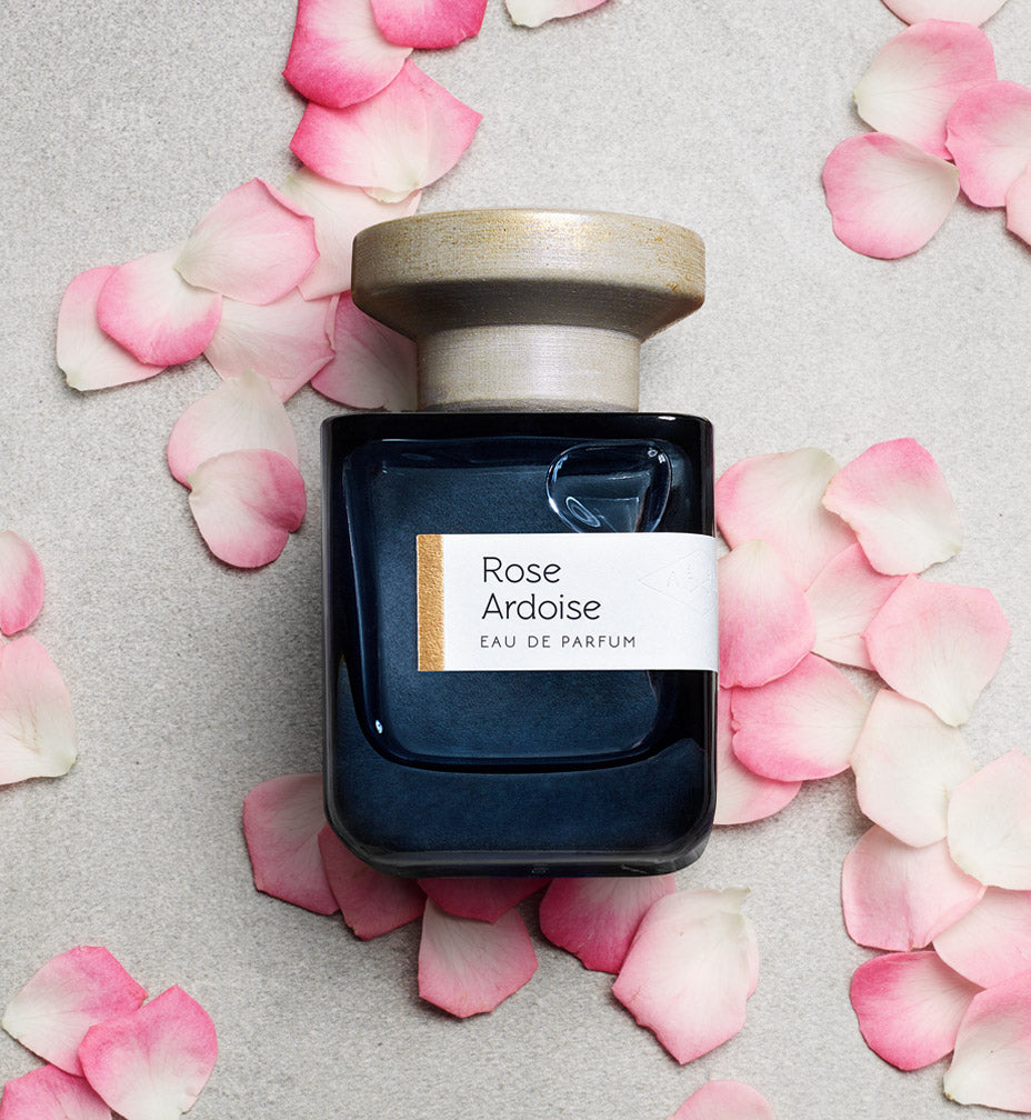 Dunkelblaue Glasflasxhe mir grauem Deckel "Rose Ardoise" von Atelier Materi, liegend, umgeben von Rosenblättern.