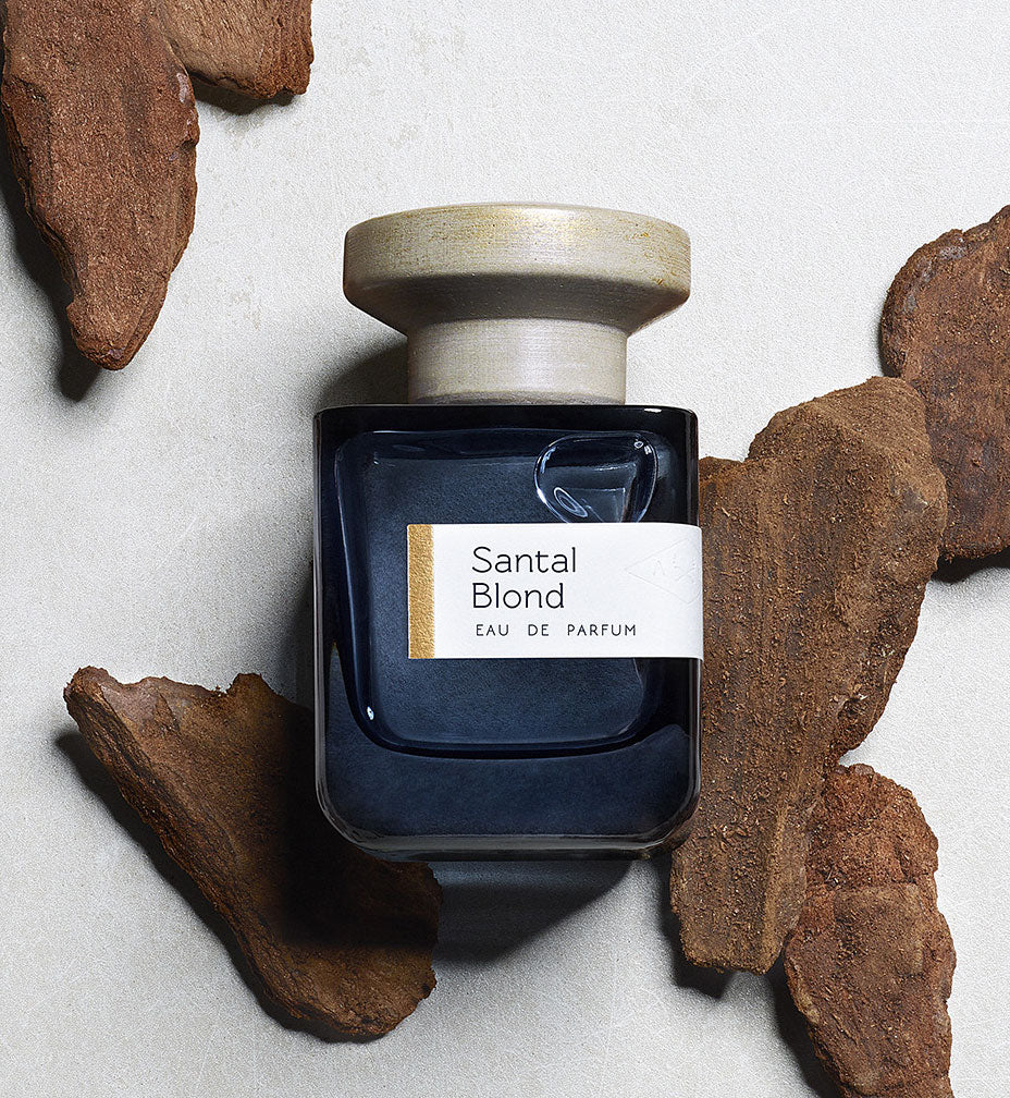 Dunkelblaue Glasflasche mit grauem Deckel  "Santal Blond" von Atelier Materi umgeben von kleinen Holzstücken.