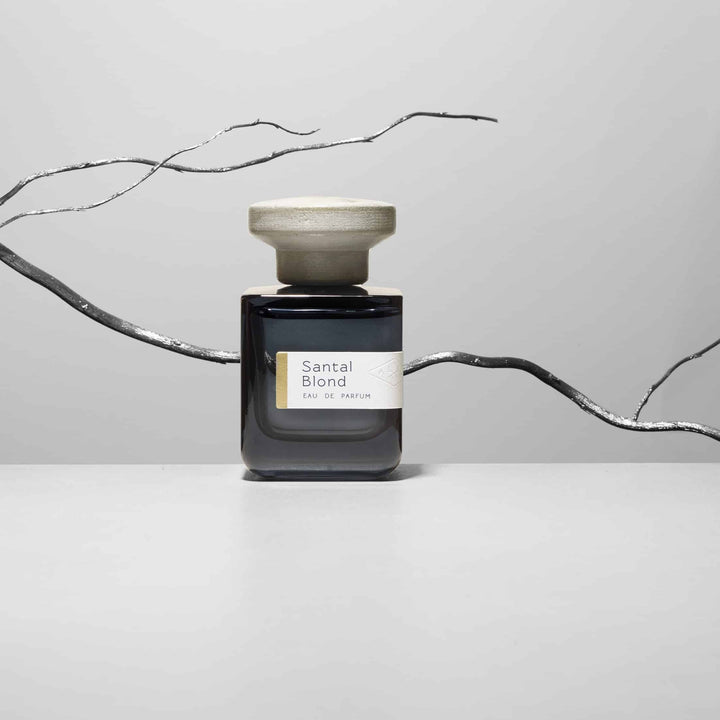 Dunkelblaue Glasflasche mit grauem Deckel  "Santal Blond" von Atelier Materi mit einem silbernen Ast im Hintergrund.