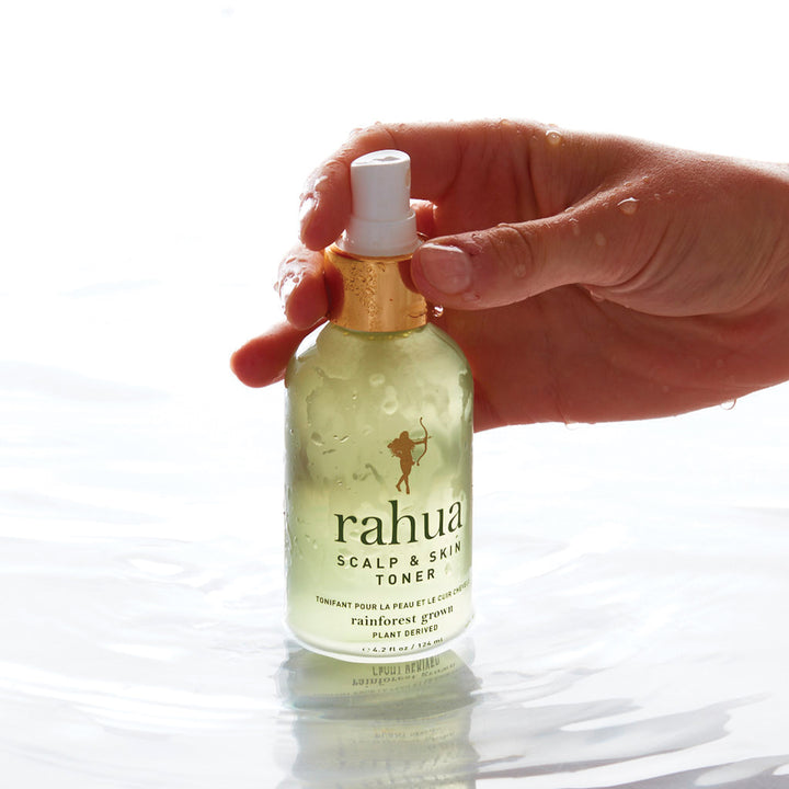 Frauenhand hält eine stehende Rahua Scalp & Skin Toner Flasche im Wasser vor hellem Hintergrund.