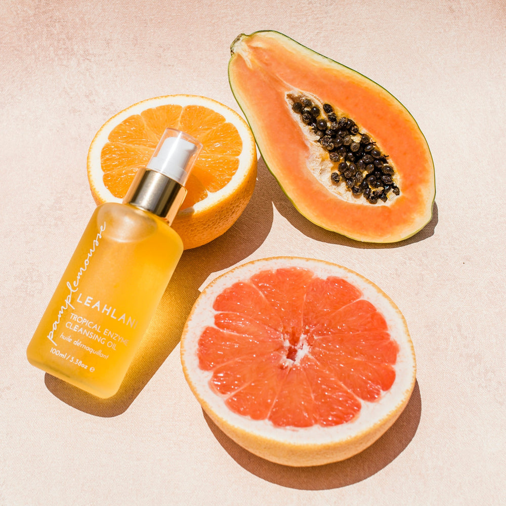 Leahlani Pamplemousse Cleansing Oil Flasche auf einer Orange mit aufgeschnittener Papaya vor hellem Hintergrund