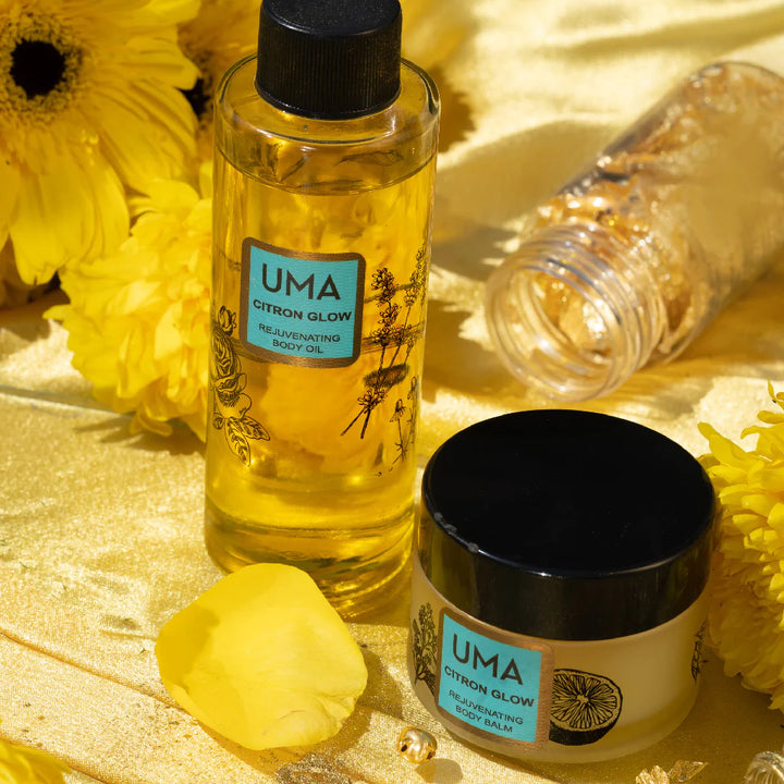 UMA Citron Glow Body Oil Flasche und Citron Glow Body Balm Schraubglas stehen auf goldener Decke zwischen gelben Blütenblättern.