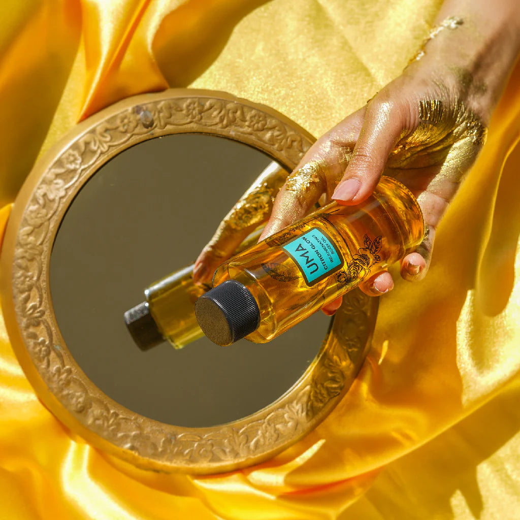 Ein antiker Spiegel liegt auf einer gelben Deckel, davor hält eine mit Gold beschmierte Hand eine UMS Cirton Glow Body Oil Flasche. North Glow