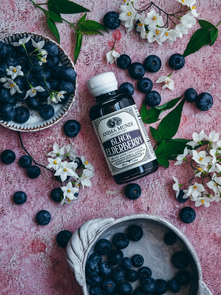Anima Mundi Glasflasche Black Elderberry aus der Vogelperspektive zwischen Holunderbeeren und -blüten.