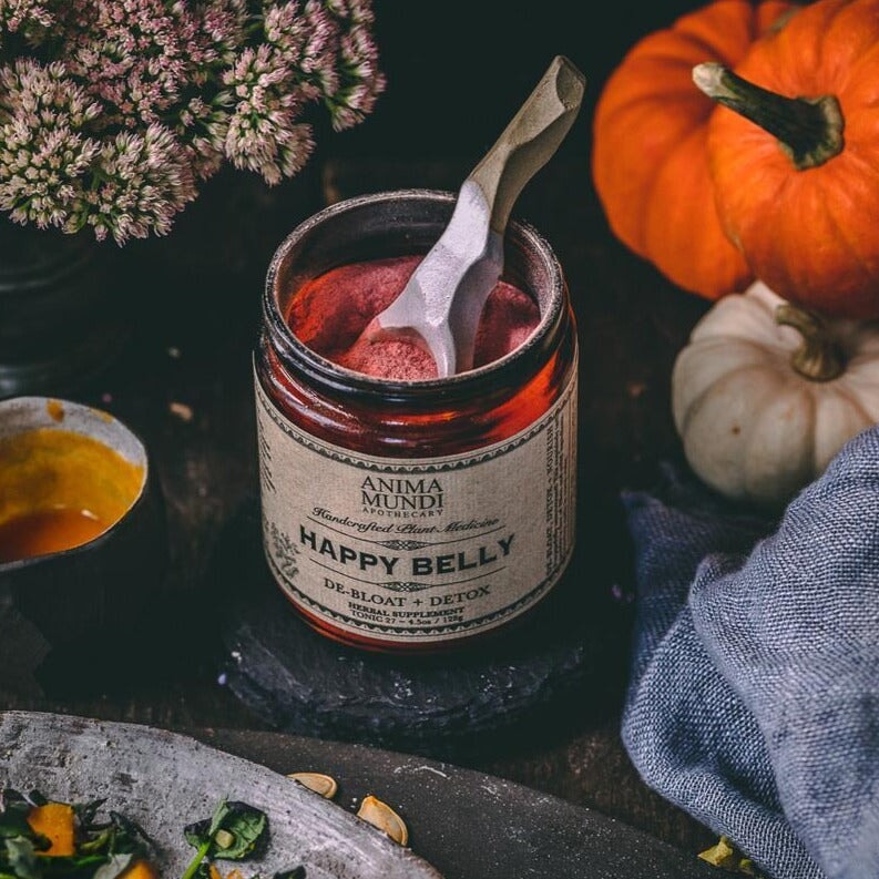 Offenes Belly Love Glas mit Holzlöffel steht zwischen Kürbissen und Blumen.