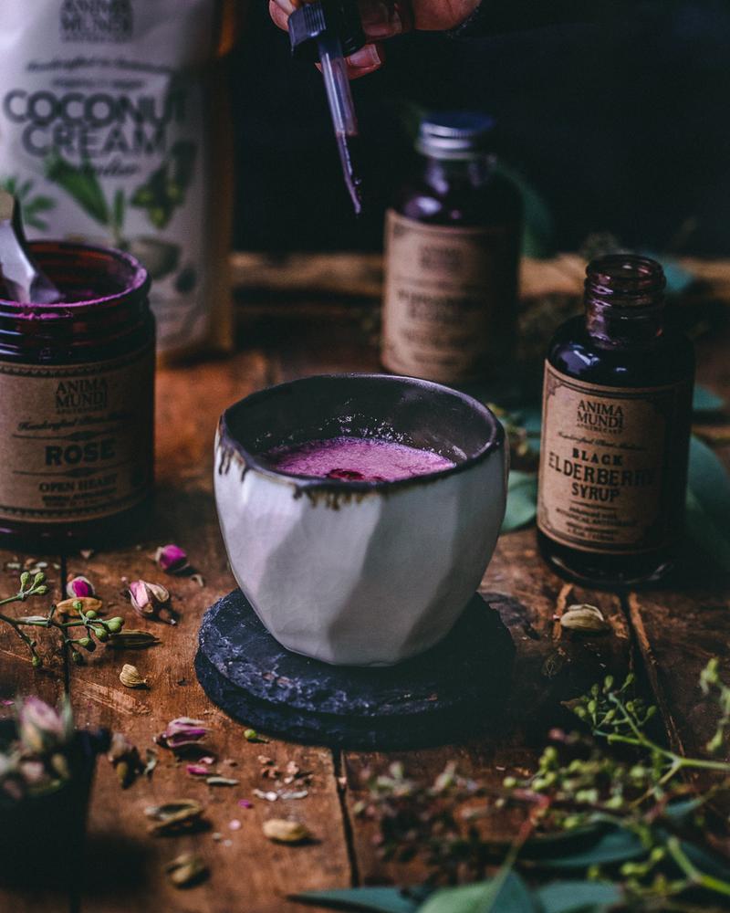 Steingutbecher mit rosafarbenem Getränk steht auf einem Holztisch vor verschiedenen Anima Mundi Produkten, eine Pipette tropft dunkellila Flüssigkeit in den Becher.
