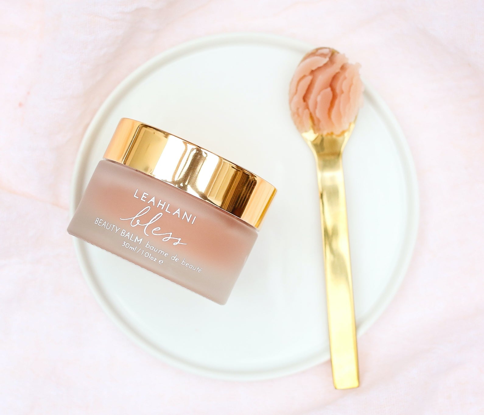 Leahlani Bless Beauty Balm Tiegel mit goldenem Deckel und goldenem Spatel mit Blam auf hellem Teller