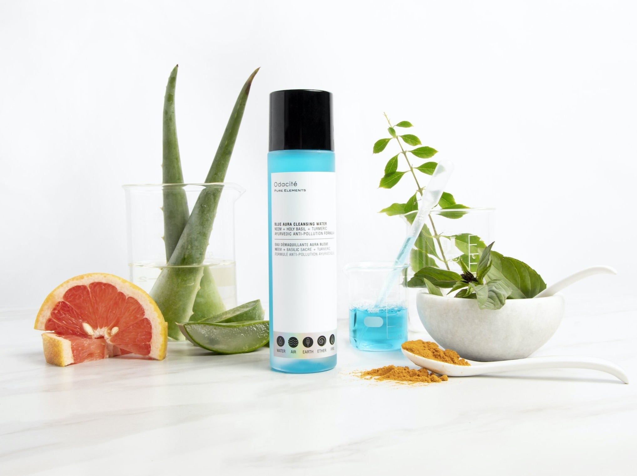 Odacité Blue Aura Cleansing Water, blaue Flüssigkeit in transparenter Flasche, arrangiert mit Aloe Vera, Grapefruit, Kurkuma und Basilikum – symbolisiert Reinheit und Clean Beauty