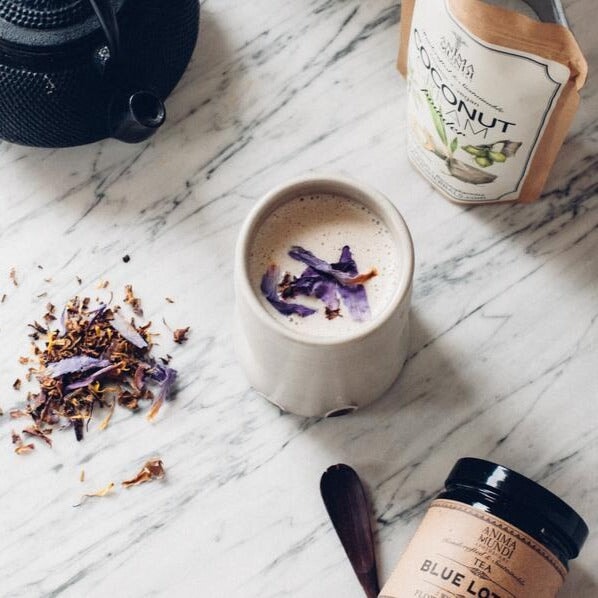 Teebecher mit milchigem Inhalt und Blue Lotus Teeblättern bestreut aus der Vogelperspektive, steht auf einem mormoriertem Tisch zwischen Anima Mundie Coconut Cream Powder, Teekanne und Blue Lotus Tee Schraubverschlussglas.