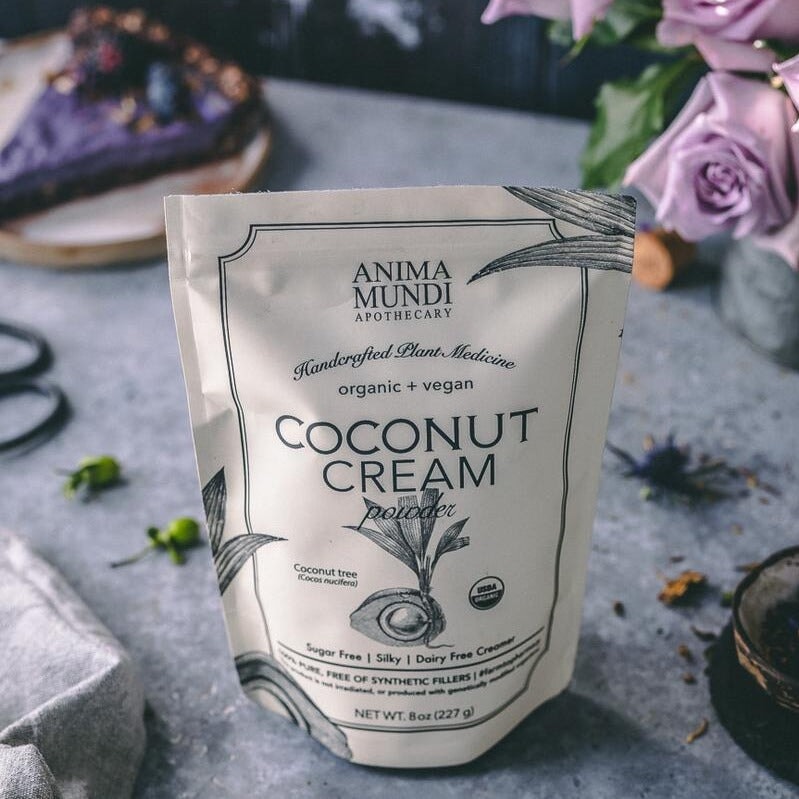 Anima Mundi Coconut Cream Powder Tüte steht zwischen Blumen und Tortenstück auf einer Steinplatte.