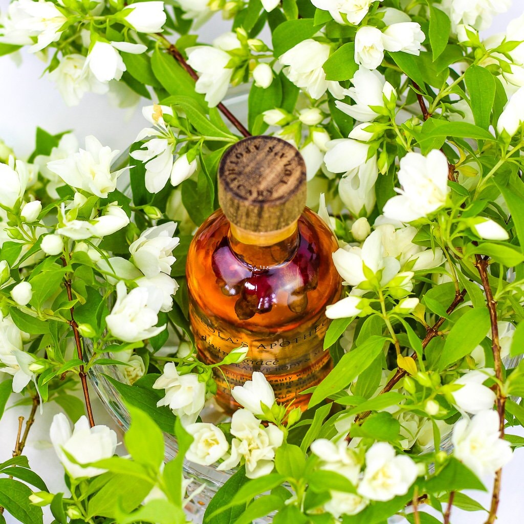 Glasflasche Lola´s Apothecary Body & Massageöl mit orangerotem Inhalt, Ansicht von oben, umgeben von Blumen.