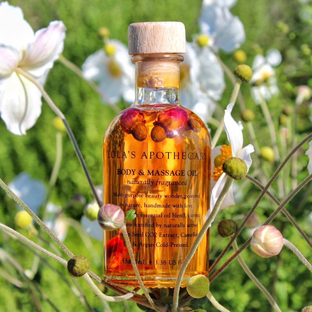 Glasflasche Lola´s Apothecary Body & Massageöl mit orangerotem Inhalt, umegeben von einer Blumenwiese.