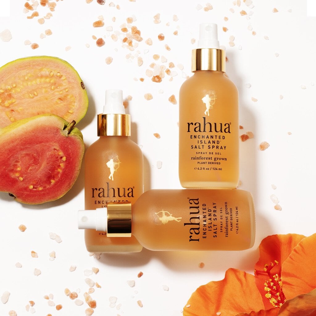 3 Rahua Enchanted Island Salt Spray Flaschen liegen auf weißer Oberfläche dekoriert mit aufgeschnittener Passionfruit, rosa Meersalzkristallen sowie Hibiskusblüte.