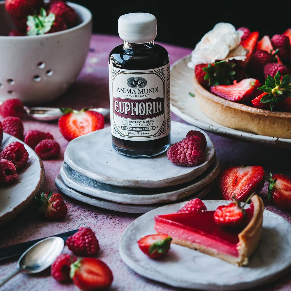 Anima Mundi Glasflasche Euphoria steht auf Steinuntersetzern dekoriert mit Erdbeeren, Himbeeren und rotem Tortenstück.